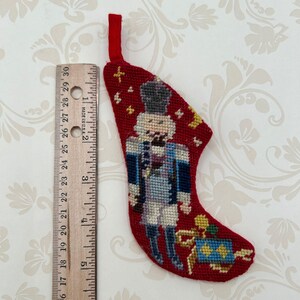 May include: A small, red Christmas stocking with a needlepoint design of a nutcracker soldier. The stocking is approximately 5 inches long and features a loop for hanging. The nutcracker is dressed in a blue and white uniform, holding a gift.