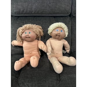 May include: Two Cabbage Patch Kids dolls. One doll has light brown hair, and the other has white hair. Both dolls have blue eyes and are wearing no clothing. The dolls are sitting on a dark gray upholstered chair.