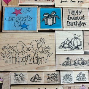May include: A collection of wooden stamps with various designs and text. Stamps include the words "congrats", "Happy Easter!", and "Happy Belated Birthday". Other stamps feature illustrations of a gift, pumpkins, and other decorative elements.