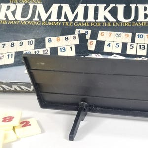 May include: A Rummikub tile game set with a black tile holder and game tiles. The game box is visible in the background, with the game's title and description printed on it. The tiles are white with black numbers.