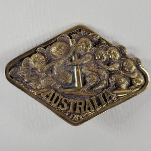 May include: A gold-tone metal belt buckle featuring a group of koalas climbing a pole with the word "AUSTRALIA" inscribed below.