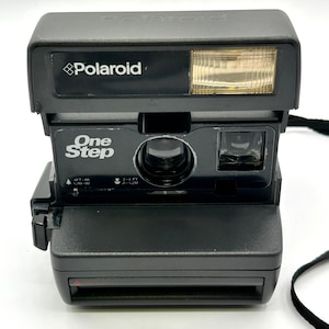 May include: A black Polaroid OneStep instant camera. The camera has a built-in flash and the Polaroid logo is visible on the top. The words "One Step" are printed on the front of the camera.