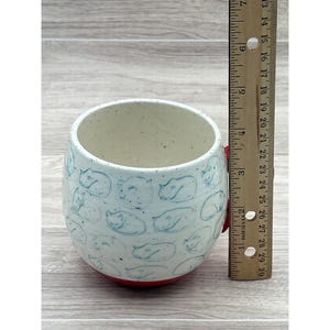May include: A ceramic mug with a white interior and exterior. The exterior is decorated with blue cat illustrations. The base is red, and the mug has a red handle. The mug is approximately 4 inches tall.