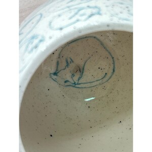 May include: A white ceramic bowl with a sleeping fox illustration in blue. The bowl's interior has a speckled appearance, and the rim features a blue, abstract design. The fox is curled up, adding a whimsical touch to the piece.