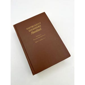 May include: A brown hardcover book titled "INSTRUMENT ENGINEERS Handbook" with gold lettering. The book is Volume 1 and covers process measurement. The author's name is also printed in gold. The book is on a white background.