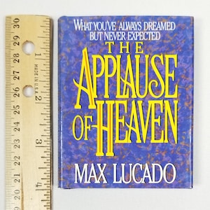 May include: A book cover with a blue background and gold lettering. The title is "The Applause of Heaven" by Max Lucado. The text on the cover reads "What you've always dreamed but never expected."