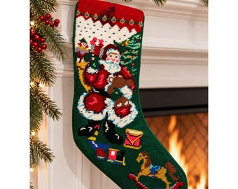May include: A green needlepoint Christmas stocking featuring a central Santa Claus figure carrying a sack of toys. The design includes a rocking horse, a toy train, and a drum on a dark green background with a white snowy border at the top.