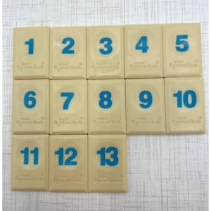 May include: A set of thirteen rectangular off-white Rummikub game tiles arranged in three rows. Each tile features a large, bold blue number ranging from one to thirteen, with the text Original Rummikub embossed below each numeral.