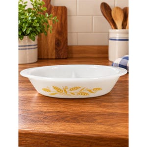 May include: A white oval divided baking dish made of milk glass, featuring a yellow wheat stalk pattern printed on the exterior side. The dish has a smooth, rounded rim and a central divider that separates the interior into two distinct sections.