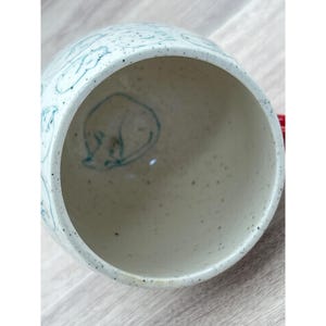 May include: A speckled ceramic mug with a light blue cat illustration inside and on the exterior. The mug has a red handle and a wide opening. The cat illustration is a simple line drawing.