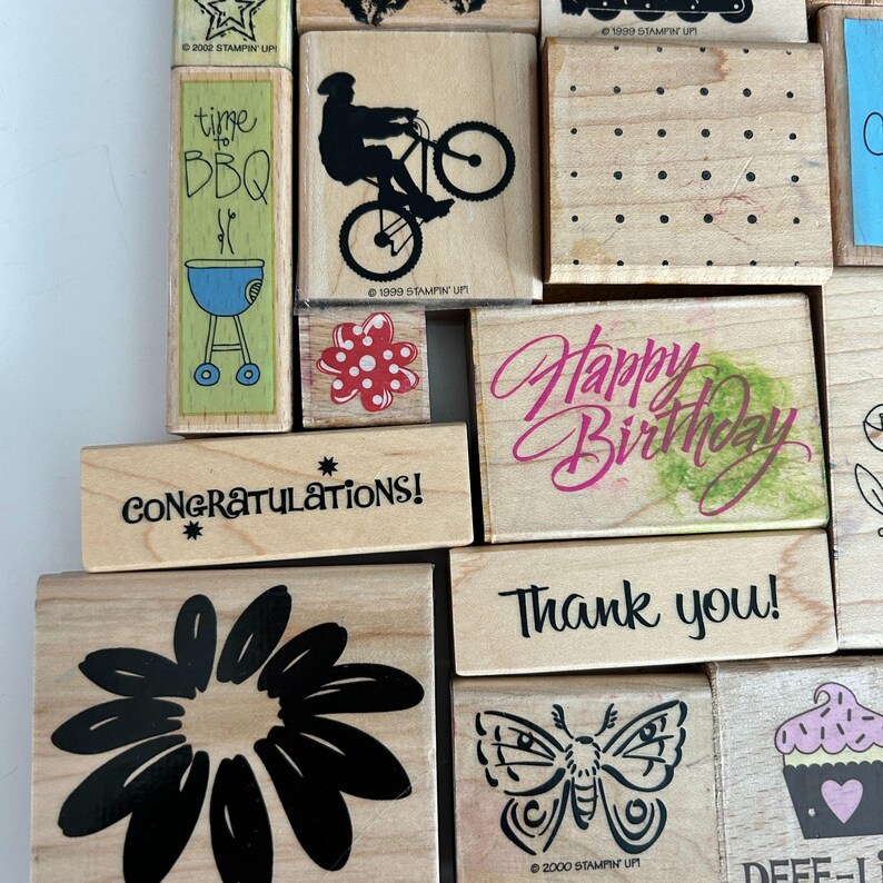 May include: A collection of wooden stamps with various designs and text. Designs include a bicycle, a daisy, a butterfly, a cupcake, and the phrases "Happy Birthday," "Thank you," and "Congratulations!" The stamps are in various sizes and shapes.