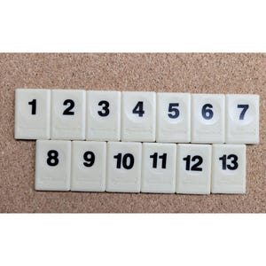 May include: A set of thirteen rectangular off-white Rummikub game tiles arranged in two rows. Each tile features a bold black number ranging from one to thirteen, with the brand name Rummikub embossed in small letters at the bottom.