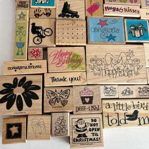 May include: A collection of wooden stamps with various designs and text. Designs include a roller skate, a bicycle rider, a flower, a butterfly, a cupcake, and a Santa Claus. Text includes "Happy Birthday," "Thank You," and "Do Not Open 'Til Christmas!"