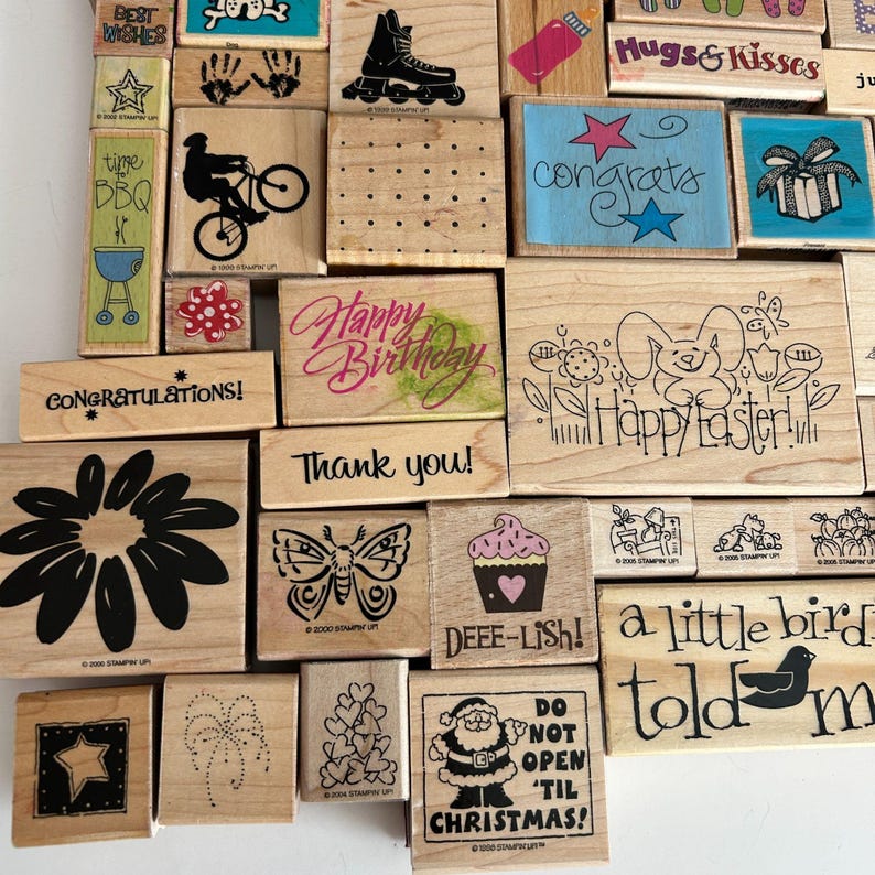 May include: A collection of wooden stamps with various designs and text. Designs include a roller skate, a bicycle rider, a flower, a butterfly, a cupcake, and a Santa Claus. Text includes "Happy Birthday," "Thank You," and "Do Not Open 'Til Christmas!"