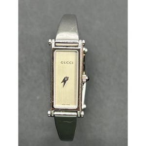 May include: A rectangular Gucci watch with a silver-tone metal band and a light-colored face. The watch face features the Gucci logo and a black arrow design. The watch has a black leather strap and a clasp closure. The words "SWISS MADE" are printed on the watch face.