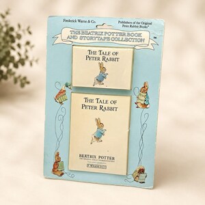 May include: A light blue cardboard display card featuring The Beatrix Potter Book and Storytape Collection. It includes two cream-colored rectangular items labeled The Tale of Peter Rabbit with illustrations of Peter Rabbit in a blue jacket.