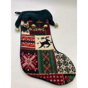 May include: A Christmas stocking with a patchwork design featuring a reindeer silhouette, snowflakes, and geometric patterns in red, green, and white. The stocking has a green velvet top with gold bells.