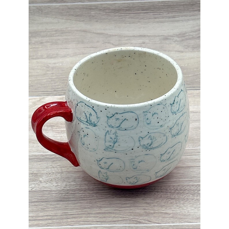 May include: A ceramic mug with a white speckled finish and a red handle and base. The mug is decorated with light blue sketches of sleeping cats. The mug is a unique and charming addition to any kitchen.