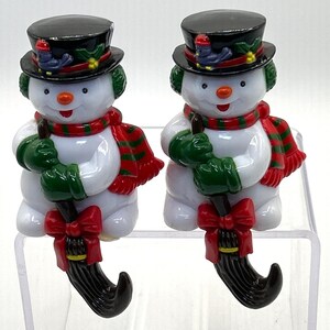 May include: Two snowman figurines. Each snowman has a black top hat, green earmuffs, a red and green scarf, green gloves, and a black broom. A red bow is at the bottom of the broom. The snowmen have orange noses and smiling faces.