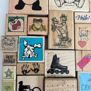 May include: A collection of wooden stamps with various designs. Images include a cartoon frog, a girl with a teddy bear, a roller skate, and the text "Hello!". Some stamps have the text "Best Wishes" and "Honey".