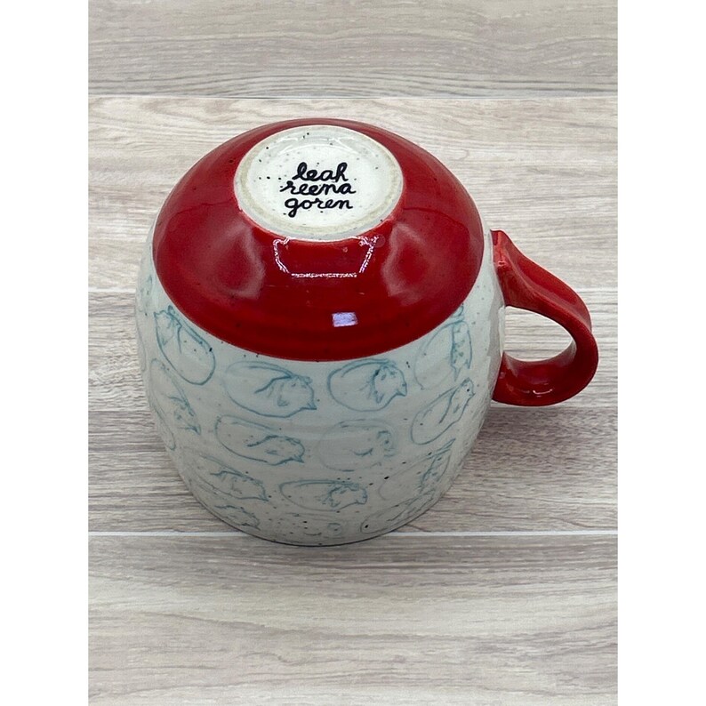 May include: A handmade ceramic mug with a red top and handle, and a white base decorated with blue line drawings. The interior base of the mug has the text "leah reena goren". The mug is designed for hot beverages.