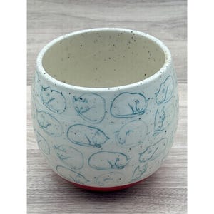 May include: A ceramic tumbler with a light blue exterior adorned with sleeping cat illustrations in a darker blue. The interior is off-white with speckles, and the base is red. This handmade cup is ideal for beverages.