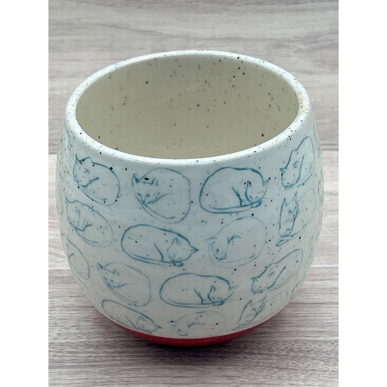 May include: A ceramic tumbler with a light blue exterior adorned with sleeping cat illustrations in a darker blue. The interior is off-white with speckles, and the base is red. This handmade cup is ideal for beverages.