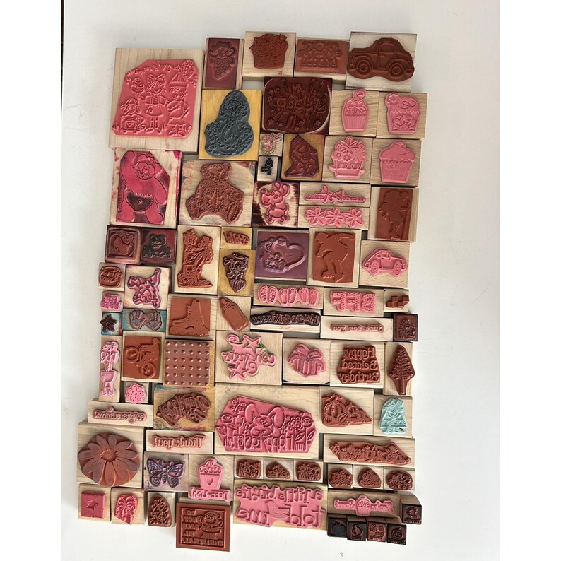 May include: A collection of various rubber stamps on wooden blocks. The stamps feature diverse designs, including animals, flowers, and text. The stamps are in shades of pink, brown, and black.