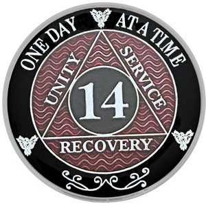 Puede incluir: Una moneda negra y plateada con un diseño de triángulo rosa. El triángulo tiene las palabras "Unidad", "Servicio" y "Recuperación" inscritas en cada lado. El número "14" está en el centro del triángulo. Las palabras "Un día a la vez" están inscritas alrededor del exterior de la moneda.