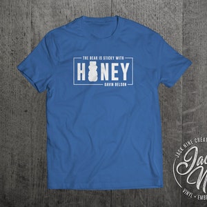 May include: A royal blue t-shirt with a white graphic that says "The bear is sticky with honey" and "Gavin Belson". The graphic also features a honey jar. The bottom right corner of the image shows a white logo that says "Jack Nine Creative" and "Jack Nine" in a stylized font. The logo also says "Vinyl, Embroidery, Apparel".
