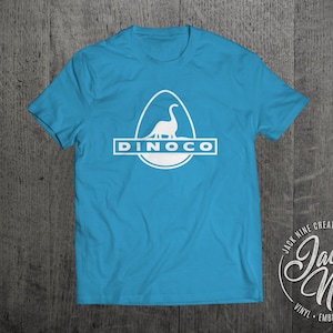 May include: A turquoise blue t-shirt featuring a white graphic of a dinosaur in an egg with the text "DINOCO" underneath. The shirt is displayed against a wooden backdrop. The Jack Nine Creative logo is in the bottom right corner.