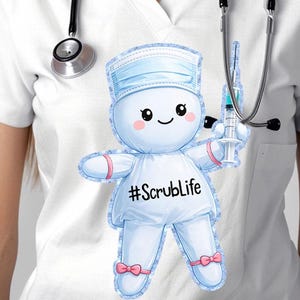May include: White V-neck scrub top with a cartoon nurse doll graphic. The doll is light blue and wearing a face mask, holding a syringe, and has the text "#ScrubLife". A black stethoscope hangs around the neck.