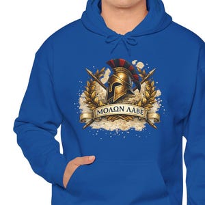 Pode incluir: Um moletom azul royal com um gráfico frontal com um capacete espartano, lanças cruzadas e a frase grega "ΜΟΛΩΝ ΛΑΒΕ" em uma faixa. O design é detalhado com detalhes dourados e vermelhos.