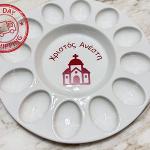 May include: A white ceramic Easter egg plate with a central well and 12 egg-shaped indentations. The center features a red church illustration and Greek text. A red "Next Day Shipping" stamp is in the upper left.