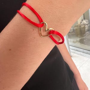 May include: A red cord bracelet featuring a gold heart-shaped charm. The bracelet is tied around the wrist. The gold heart charm is the focal point of the bracelet.