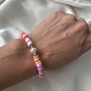 Op de afbeelding: Een kleurrijke armband met platte, ronde kralen in verschillende kleuren, waaronder roze, geel, paars en groen. De armband heeft een parel en gouden accenten. De armband zit om een pols.