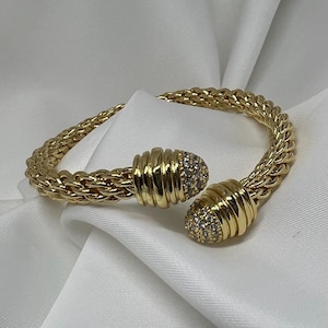 Può includere: Un bracciale a polsino dorato con un motivo a catena intrecciata. Il bracciale presenta due estremità decorative con una texture a coste ed è incrostato di piccole pietre preziose trasparenti. Il bracciale è esposto su uno sfondo di tessuto bianco.