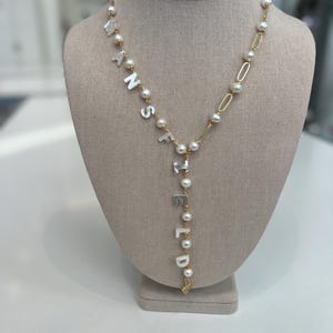 May include: A gold-tone lariat necklace featuring white mother-of-pearl letter beads spelling MANSFIELD, interspersed with round white pearls and gold-tone chain links. The design includes a vertical drop section and a small gold-tone charm at the end.