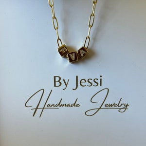 Puede incluir: Collar de cadena dorada con un colgante que deletrea el nombre "RVA" en letras doradas en bloque con pequeños diamantes. El collar está sobre un fondo blanco con el texto "By Jessi Handmade Jewelry".