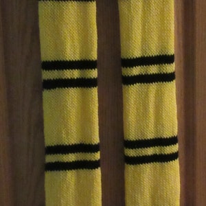 May include: A yellow and black knitted scarf with black stripes. The scarf has a fringe at the bottom. The scarf is hanging on a hook against a wooden door.