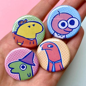 Pode incluir: Quatro botões coloridos de personagens de desenhos animados com fundo de bolinhas. Os personagens são uma criatura amarela com um chapéu, uma criatura rosa com olhos grandes, uma criatura vermelha com uma gravata e uma criatura verde com um chapéu.