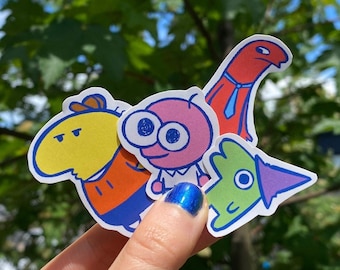May include: Four colorful cartoon character stickers. The stickers are yellow, pink, green, and red. The stickers are on a hand.