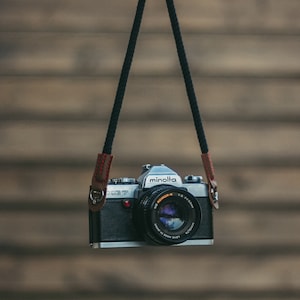 May include: A black camera strap with brown leather accents is hanging from a hand. A silver camera with a black lens is attached to the strap. The camera has the word "minolta" printed on it.