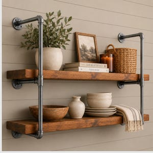 May include: A two-tier wall-mounted shelving unit featuring dark stained wooden planks supported by dark gray industrial metal pipes. The shelves hold a potted plant, a framed picture, books, a woven basket, a candle, bowls, and a striped cloth.