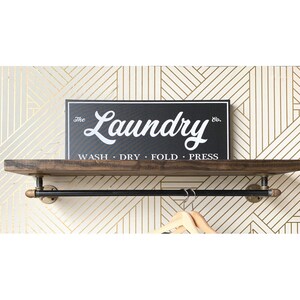 May include: A wall-mounted shelf made of dark brown wood with a black metal pipe hanging rod underneath. A rectangular black sign with white text reads The Laundry Co. WASH &middot; DRY &middot; FOLD &middot; PRESS. Two light-colored wooden clothes hangers are visible.