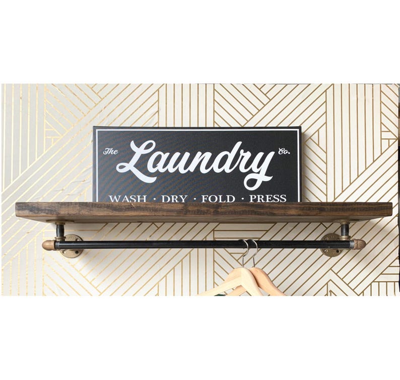 May include: A wall-mounted shelf made of dark brown wood with a black metal pipe hanging rod underneath. A rectangular black sign with white text reads The Laundry Co. WASH &middot; DRY &middot; FOLD &middot; PRESS. Two light-colored wooden clothes hangers are visible.