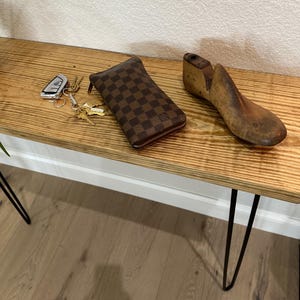 May include: A brown checkered wallet with a zipper closure, keys, and a vintage wooden shoe last rest on a wooden table. The table has black hairpin legs and is against a white wall.