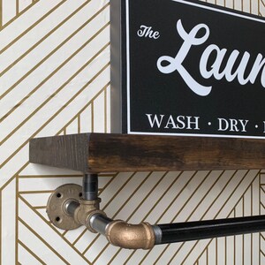 May include: A dark brown wooden shelf mounted on industrial-style metal pipe brackets with a black horizontal bar. A black rectangular sign sits on the shelf with white text that reads The Laundry, WASH, DRY, FOLD in a script and sans-serif font.