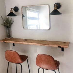 Pode incluir: Uma configura&ccedil;&atilde;o de penteadeira r&uacute;stica com um espelho retangular com moldura dourada, duas arandelas pretas com detalhes em lat&atilde;o e uma prateleira de madeira natural. Duas banquetas de bar de couro marrom com pernas de metal preto est&atilde;o posicionadas abaixo.