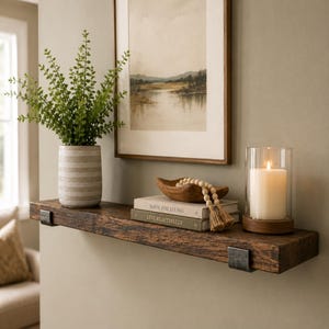 May include: A rustic dark wood floating shelf with metal brackets holding a gray patterned vase with a green plant, a wooden bowl, two stacked books titled Made for Living and Live Beautifully, wooden beads, and a white candle in a glass holder.